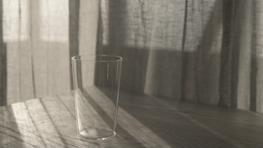 Slow motion thin tumbler glass on white oak table with curtains sways on the wind on background with sunset light | Shutterstock HD Video #1091771483