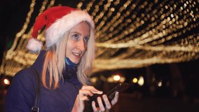 Portrait of young beautiful blonde in Santa Claus hat, typing message on mobile phone and emotionally reacting to receiving good news. looks into smartphone against background of night city lights.