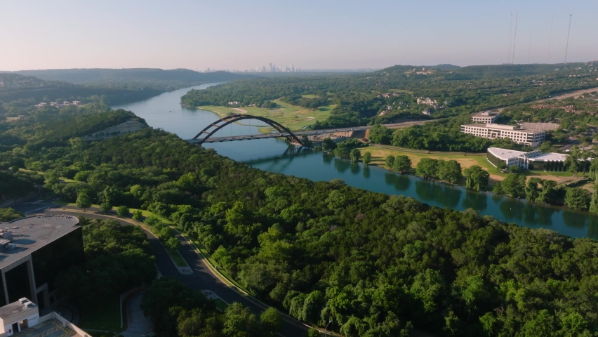 Aerial sweep of the iconic Pennybacker 360 bridge in Austin, Texas during hazy summer sunrise morning with downtown skyline in backdrop. Flying over lake and forest trail with 4k drone in 2022 Royalty-Free Stock Footage #1091773877