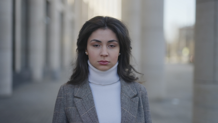 Portrait of a young woman with dark hair in a jacket looks seriously at the camera then smiles. Against the backdrop of an office building. Slow motion 4k footage