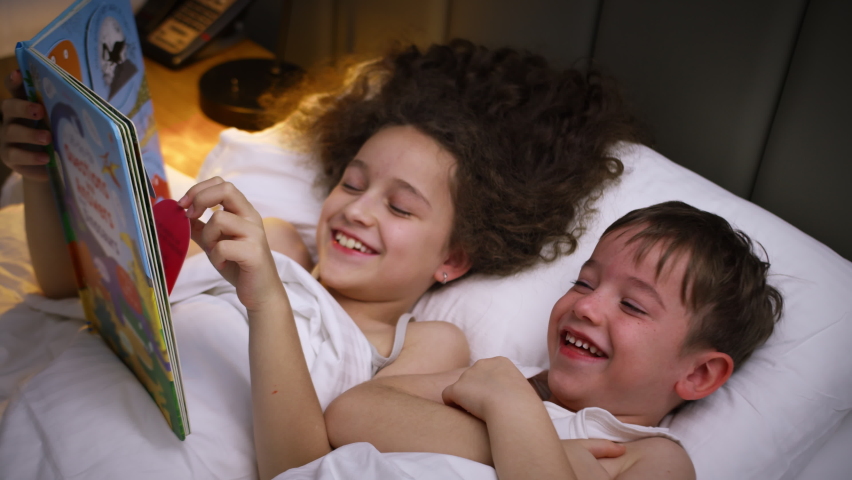 Child care at home, little sister takes care of younger brother, reads a bedtime story to children, Leisure time for children. Daughter reading a book to her brother while lying in bed at home | Shutterstock HD Video #1091779107
