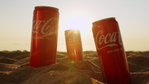 Cold cans Coca Cola sandy beach with sunlight background. man's hand takes a can of fresh Cola from a sandy beach. Close-up of three cans cola ocean. Gdansk, Poland. 06.30.2022.