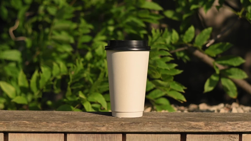 A white paper coffee tea cup with black plastic lid sits on wooden bench in the park. Hot drink to go, takeaway coffee tea. Empty space for your logo, mockup. Blurred background | Shutterstock HD Video #1091791065