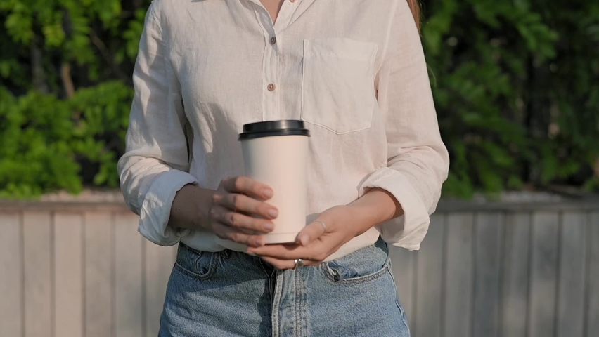 Girl in white linen shirt and blue shorts holds out paper coffee cup in the park. Paper glass of takeaway coffee tea in hands close up. Woman is enjoying drink, walk in green park mockup | Shutterstock HD Video #1091791073