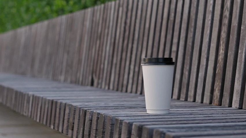 A white paper coffee tea cup with black plastic lid sits on wooden bench in the park. Hot drink to go, takeaway coffee tea. Empty space for your logo, mockup. Blurred background | Shutterstock HD Video #1091791075
