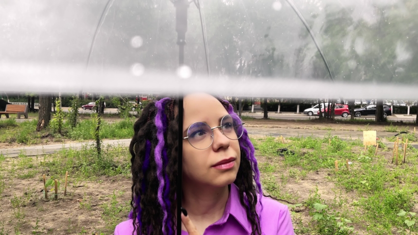A woman with umbrella in hand in the rain. Girl with glasses under an umbrella against the backdrop of an urban landscape. Female with dreadlocks stands in a park with an umbrella