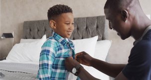 Video of happy african american father dressing up son in bedroom. Family, spending quality time together at home concept.