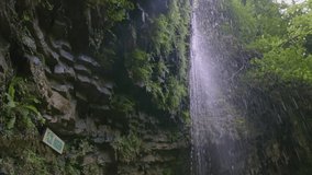 A stream of water slowly flows down the rocks. CREATIVE. A woman is standing under a waterfall. The camera from top to bottom shows a waterfall and a girl near it