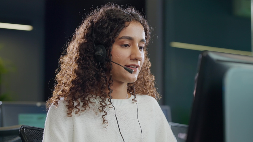 A modern attractive smiling young Indian Asian call center or customer care support curly haired female or woman employee in headset mic busy talking or interacting with client in a corporate office. Royalty-Free Stock Footage #1091806251
