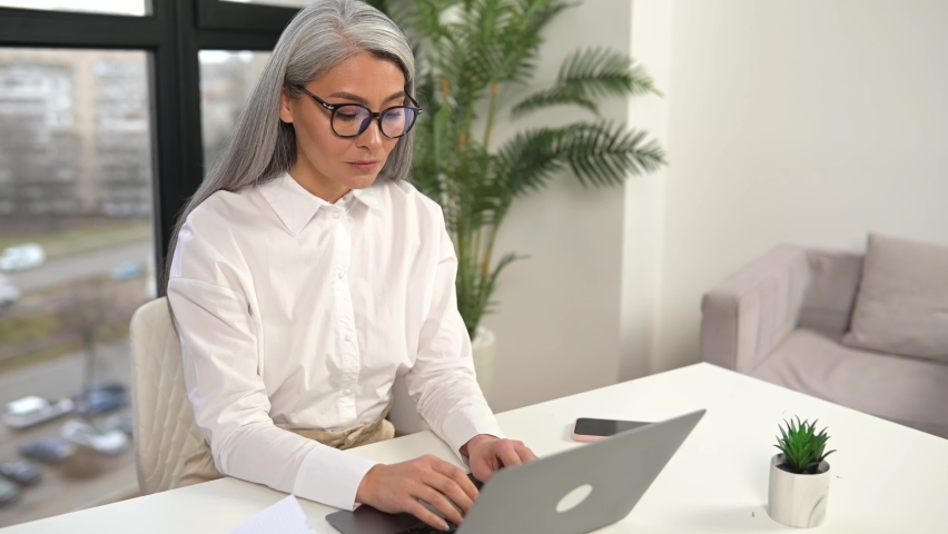 Tired mature female employee feel bored. Upset senior gray-haired woman sitting in front of the laptop, leans head on the hand, feeling headache and burnout | Shutterstock HD Video #1091806525