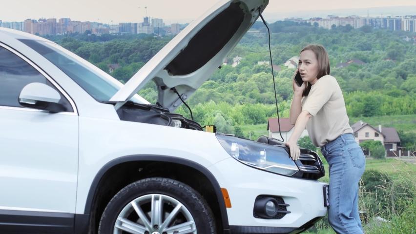 Young female driver talking on mobile phone standing near her car with open hood having engine problems | Shutterstock HD Video #1091811935