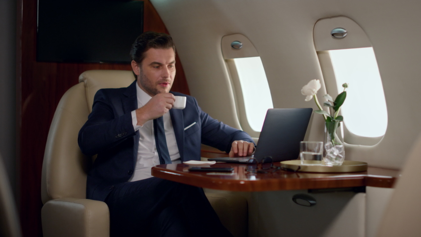 Finance manager take sip of coffee on business trip. Confident boss work laptop checking marketing sales statistics on luxury private jet. Focused stylish man looking airplane window drinking beverage