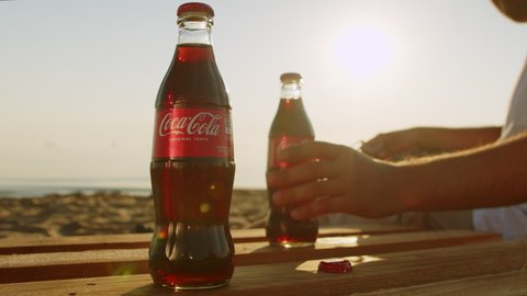 Man on a gorgeous beach in Oregon, opens a bottle of Coca cola at a beautiful sunset. 30.06.2022. young man drinks Coca-Cola Classic sandy beach. Drink is made and manufactured by Coca-Cola Company.