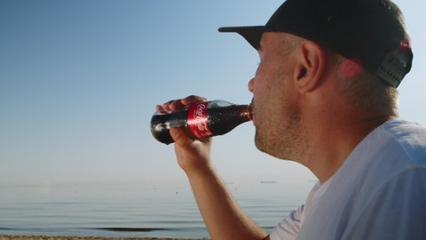 Young man drinks Coca-Cola Classic on a sandy beach. Man in a cap drinks coca-cola and is happy to cool off at a summer resort while relaxing on the beach. SANTORINI-GREECE 30.06.2022.