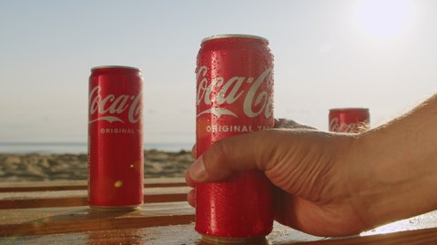 Three cans of cold Coca Cola on the seashore at sunset with the sandy beach of Gdnsk. Poland. 06.30.2022. Close-up man hand taking can of coca cola while relaxing on the beach with beautiful sunsets