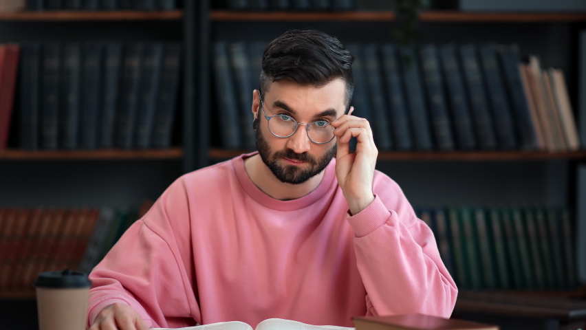 Portrait modern Hispanic male student smiling posing at library desk reading book with positive emotion. Happy hipster guy in pink sweatshirt enjoying break prepare to university exam with textbook | Shutterstock HD Video #1091823523
