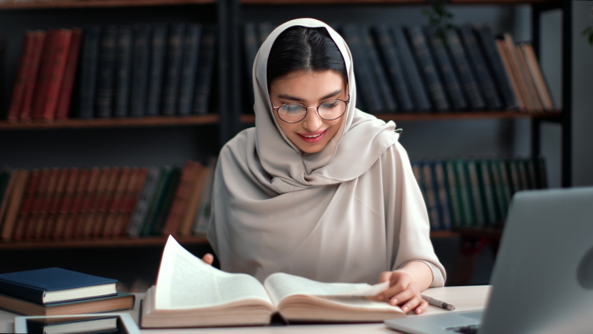 Muslim woman in hijab reading book prepare to exam at public library desk with laptop. Smiling mixed race female student teacher learning textbook search information for lecture at college university  - Powered by Shutterstock - Get 15% off with code: PIKWIZARD15