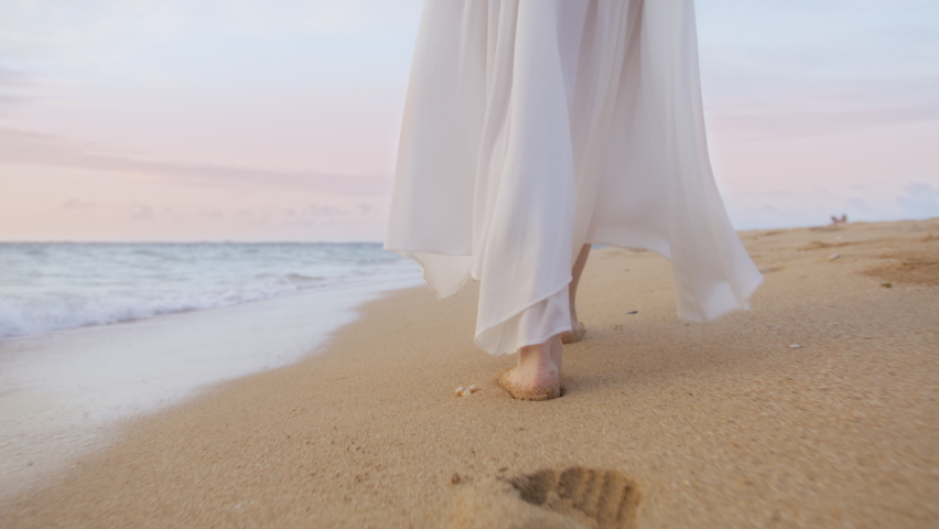 Female on romantic summer vacation island. Close up woman feet walking barefoot by beach in sunset light. Slow motion model in white summer beach dress leaving footprints in wet sand and sea waves Royalty-Free Stock Footage #1091828149