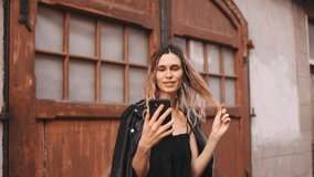 Attractive blonde curly woman with two thin braids in black clothes recording travel video blog on the old street she say hello, wave her hand, start telling, curl her hair on finger. Selective focus.