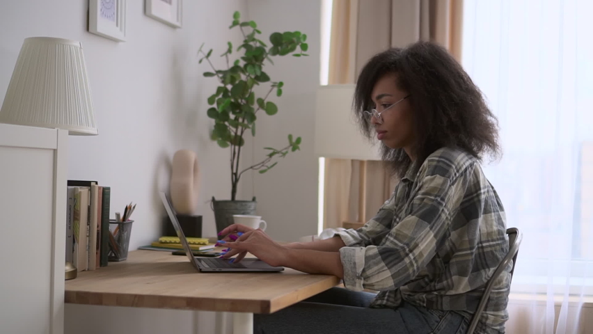 Young woman is using laptop for online work and sitting at table in home room spbd. Side view of american african american female looks at computer screen and types test, works or studies with smile | Shutterstock HD Video #1091833421