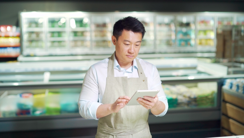 portrait of handsome asian staff man salesman in apron standing and looking at camera in grocery store supermarket. Male worker seller or small business owner. indoor. food, groceries market smile Royalty-Free Stock Footage #1091836291