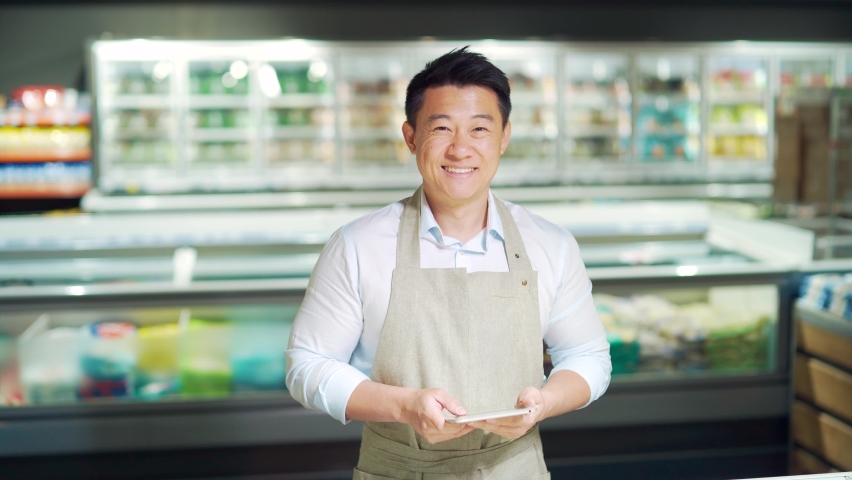 Portrait of handsome asian staff man salesman in apron standing and looking at camera in grocery store supermarket. Male worker seller or small business owner. indoor. food, groceries market smile | Shutterstock HD Video #1091836291