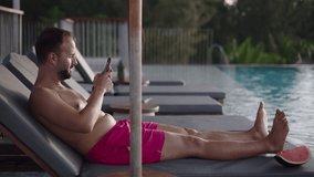 Side view of a bearded mid-age man resting on a deckchair by the pool and taking panoramic photo with a mobile phone at sunset