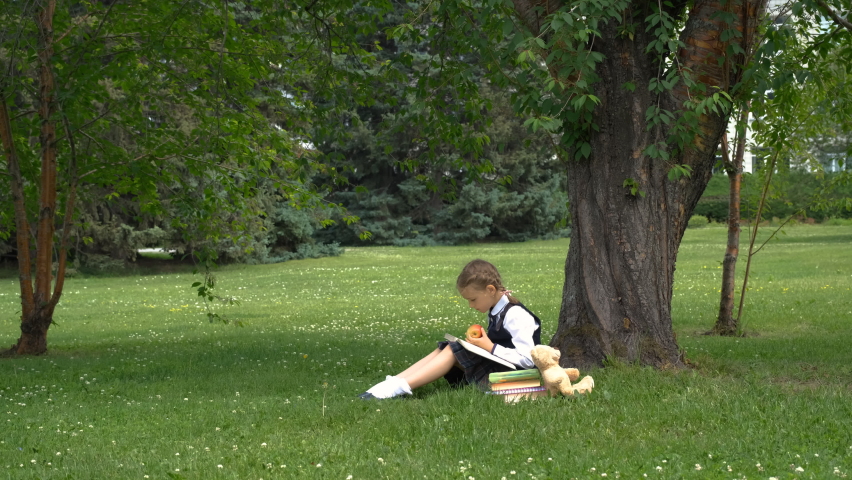 Little girl in school uniform reading a book while sitting on grass in a park. Literature, education and back to school concept | Shutterstock HD Video #1091844303