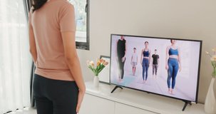 Young attractive Asian woman in sportswear site on mat in the floor virtual fitness class with group of people on a video conference on television practice yoga for beginner in living room at home.