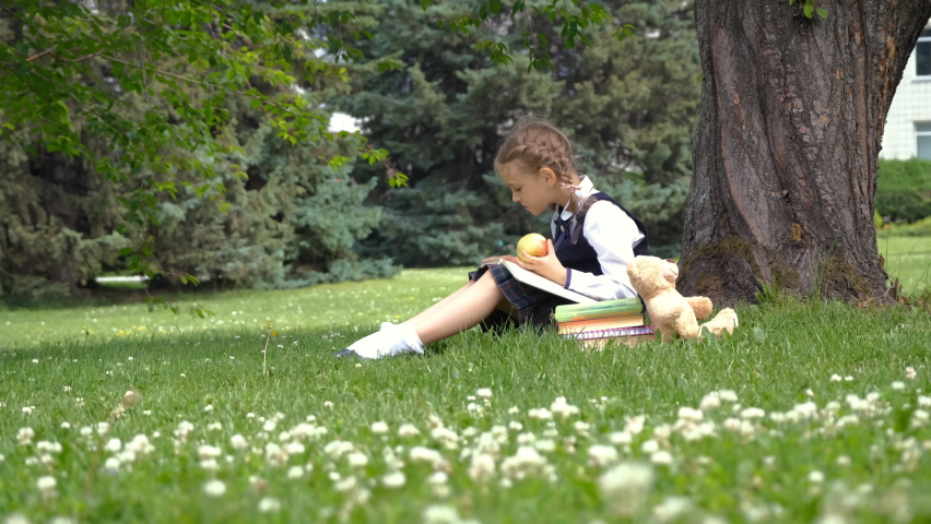 Little girl in school uniform reading book and eating fresh apple outdoors. Leisure, literature and education concept | Shutterstock HD Video #1091856093