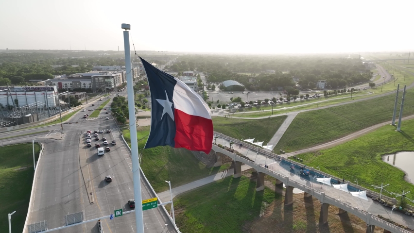 Texas Flag waves in wind. Aerial orbit reveals Trinity River and downtown Dallas TX skyline. Royalty-Free Stock Footage #1091856597