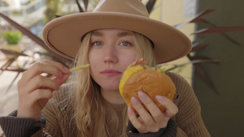 Pretty woman eating burger and French fries at the city cafe. Portrait of a young, caucasian lady enjoying her meal outdoors. High quality 4k footage | Shutterstock HD Video #1091859977