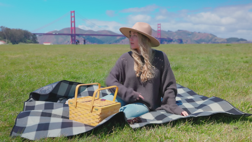 Caucasian woman sitting on a grass with the Golden Gate Bridge in the background. Cheerful fair-haired girl waving someone in the park. High quality 4k footage | Shutterstock HD Video #1091860005