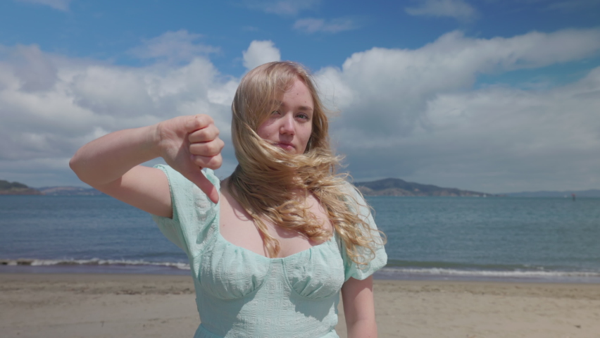 Caucasian woman thumbs down for idyllic coastal landscape. Blonde lady in bluish dress isn't happy with the weather at the beach. High quality 4k footage | Shutterstock HD Video #1091860007