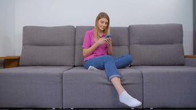 Beautiful Ukrainian female typing message on mobile phone while sitting on couch at home. Cheerful blonde woman communicating online with modern blue smartphone gadget