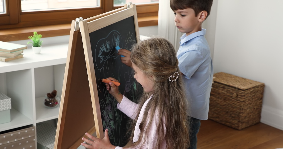 Two little sibling boy and girl drawing in chalk on play blackboard, using crayons, hatching doodles, enjoying playtime, study activity, creative art hobby in playroom. Childhood concept Royalty-Free Stock Footage #1091860841