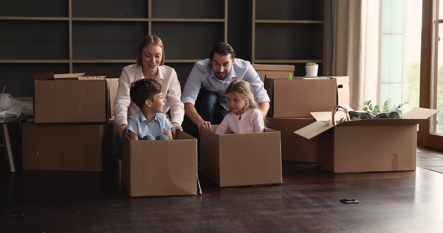 Excited little kids and parents playing racing, moving into new home. Happy young family couple dragging cardboard boxes with sibling children inside on floor, playing active game, having fun Royalty-Free Stock Footage #1091860853