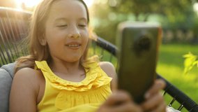 Happy kid girl holding smartphone having video call with friend distantly, online talk in the park outdoor, child using smartphone at home garden, backyard, sunlight, communication, technology concept