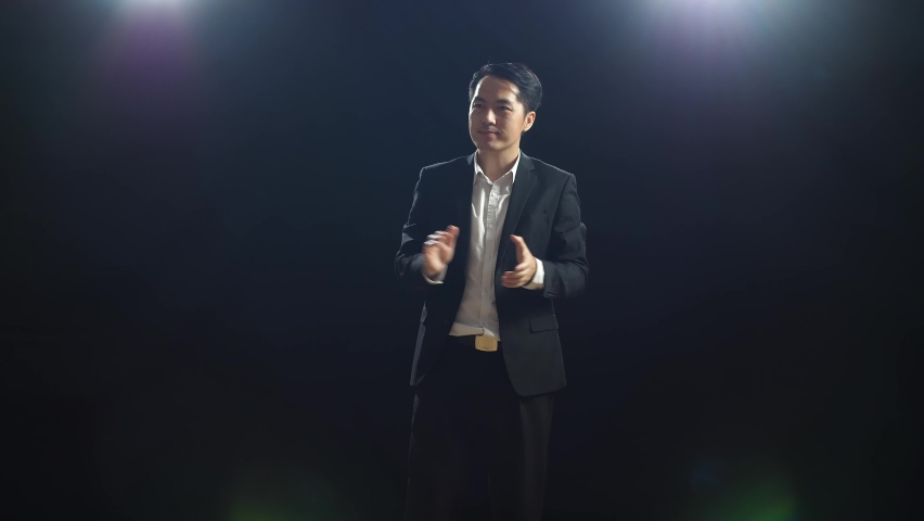 Smiling Asian Speaker Man In Business Suit Clapping His Hands And Looking Around While Standing In The Black Screen Studio
