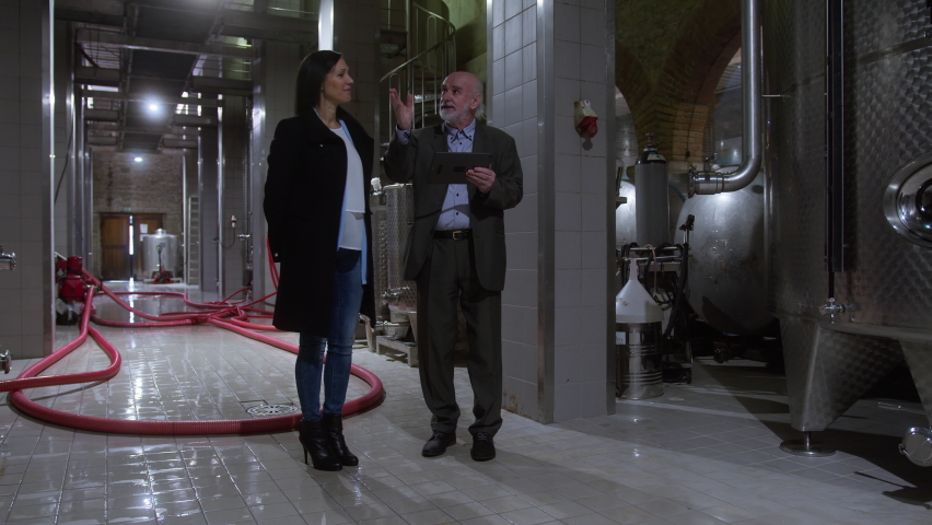 Senior vintner guiding a female visitor through his winery, explaining the process and equipment in wine industry at production cellar by using tablet, wide shot.