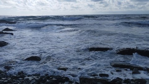The frothy sea wave hits the rocky shore and the concrete blocks lying on the shore and splashes around. The sky above the sea is blue with clouds that cover the sun