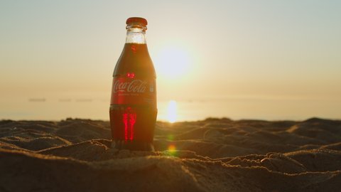 Coca-cola bottles on the sandy beach on the sunset. close-up man's hand takes a cold bottle of coca-cola on the ocean. Delete Coca Cola soft drink on the sandy beach of Gdansk. Poland. 06.30.2022.