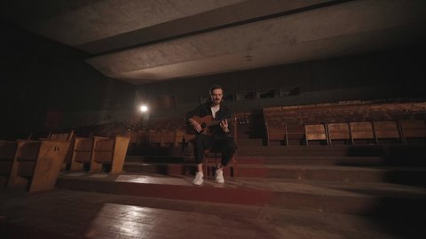 KYIV, UKRAINE - MAY 2022: A handsome guy plays an acoustic guitar in an abandoned cinema. The musician sings a song and accompanies on the guitar