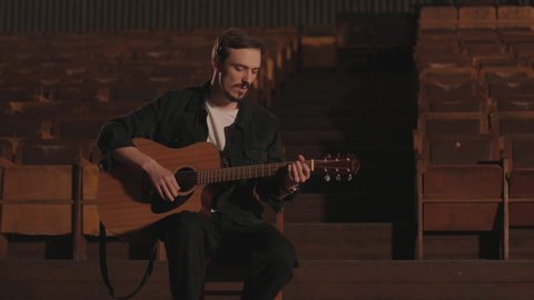 A handsome guy plays an acoustic guitar in an abandoned cinema. The musician sings a song and accompanies on the guitar