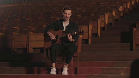 KYIV, UKRAINE - MAY 2022: A handsome guy plays an acoustic guitar in an abandoned cinema. The musician sings a song and accompanies on the guitar