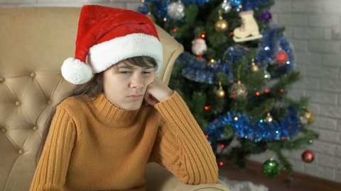 Unhappy teen by Christmas tree. A sad teen girl in Santa hat sits in loneliness in armchair by Christmas tree.
