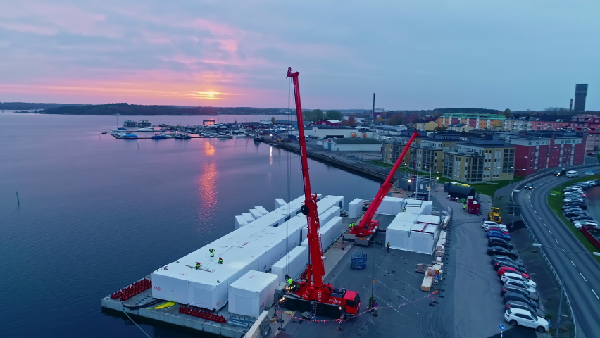 Aerial drone shot of shipping containers being loaded onto a container ship along seaside with sun going down in the background.