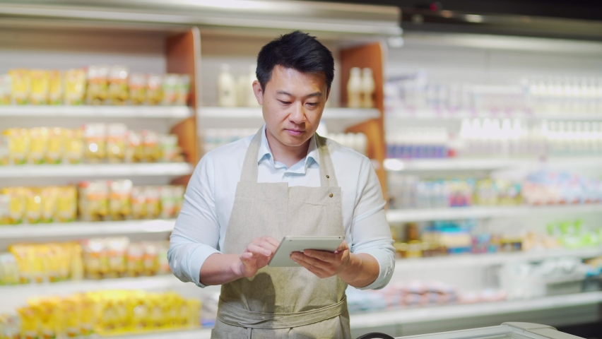Portrait of happy asian male small business owner standing in grocery store behind counter. Male manager with a tablet looking at the camera. friendly administrator food market. worker in apron | Shutterstock HD Video #1091889445