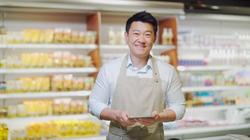 Portrait of happy asian male small business owner standing in grocery store behind counter. Male manager with a tablet looking at the camera. friendly administrator food market. worker in apron | Shutterstock HD Video #1091889445