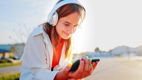 Happy teenage girl with headphones holds phone in her hands and watches video with smile. Using social networks and modern technologies for communication and entertainment. Summer leisure in nature. 